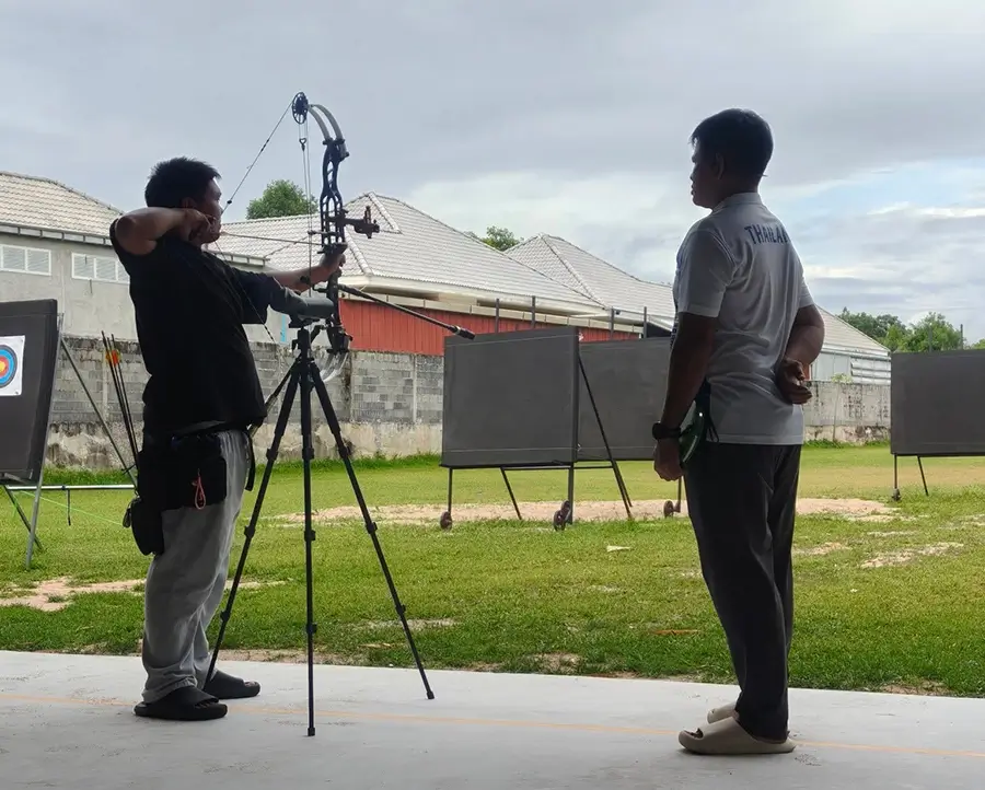 A student aims at a target in archery, guided by a coach, as another man captures the action with a camera.